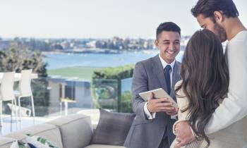 Un couple enlacé sur une terrasse dans un environnement luxueux échange avec un agent immobilier souriant, qui tient une tablette dans les mains 