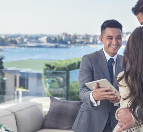 Un couple enlacé sur une terrasse dans un environnement luxueux échange avec un agent immobilier souriant, qui tient une tablette dans les mains 